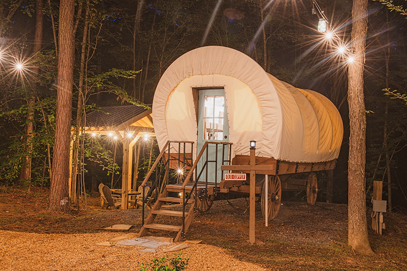 A cozy glamping setup with a covered wagon glowing warmly in a wooded area at night. A small staircase leads to its door, while string lights and a nearby gazebo with seating create a charming, secluded retreat