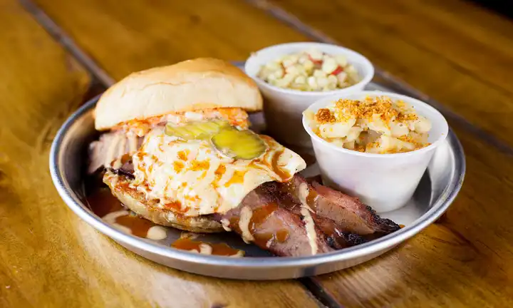 A mouthwatering Edley's BBQ plate featuring a brisket sandwich with egg, pickles, BBQ sauce, creamy mac & cheese, and Southern corn salad on a metal tray.