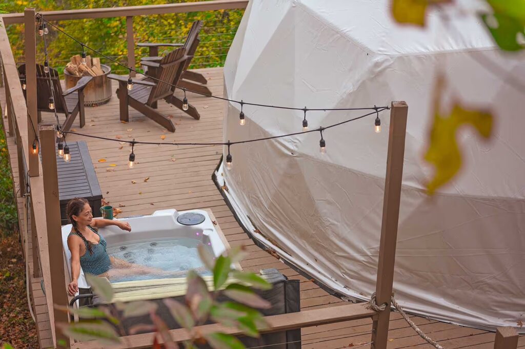 A woman relaxes in a hot tub on a cozy deck next to a dome tent, surrounded by nature—an ideal Mother’s Day escape in a peaceful forest setting.