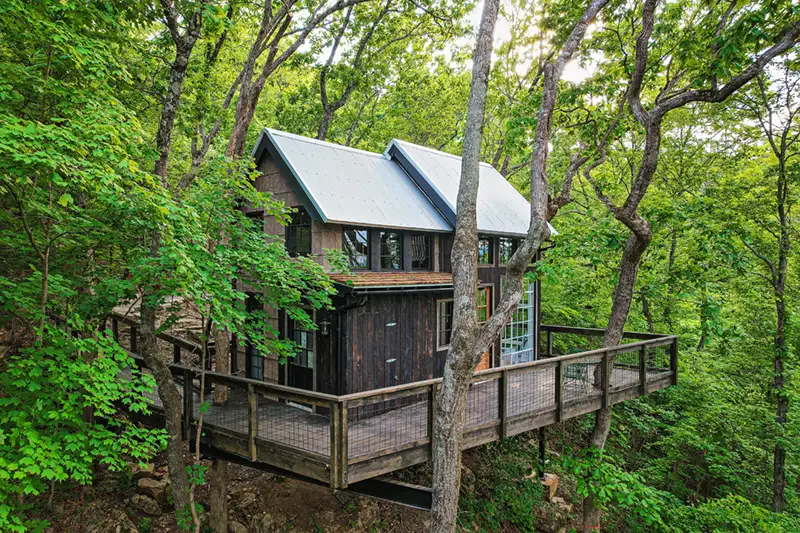 Modern treehouse cabin in Chattanooga, TN, nestled in lush woods with a wraparound deck, metal roof, and large windows surrounded by greenery.