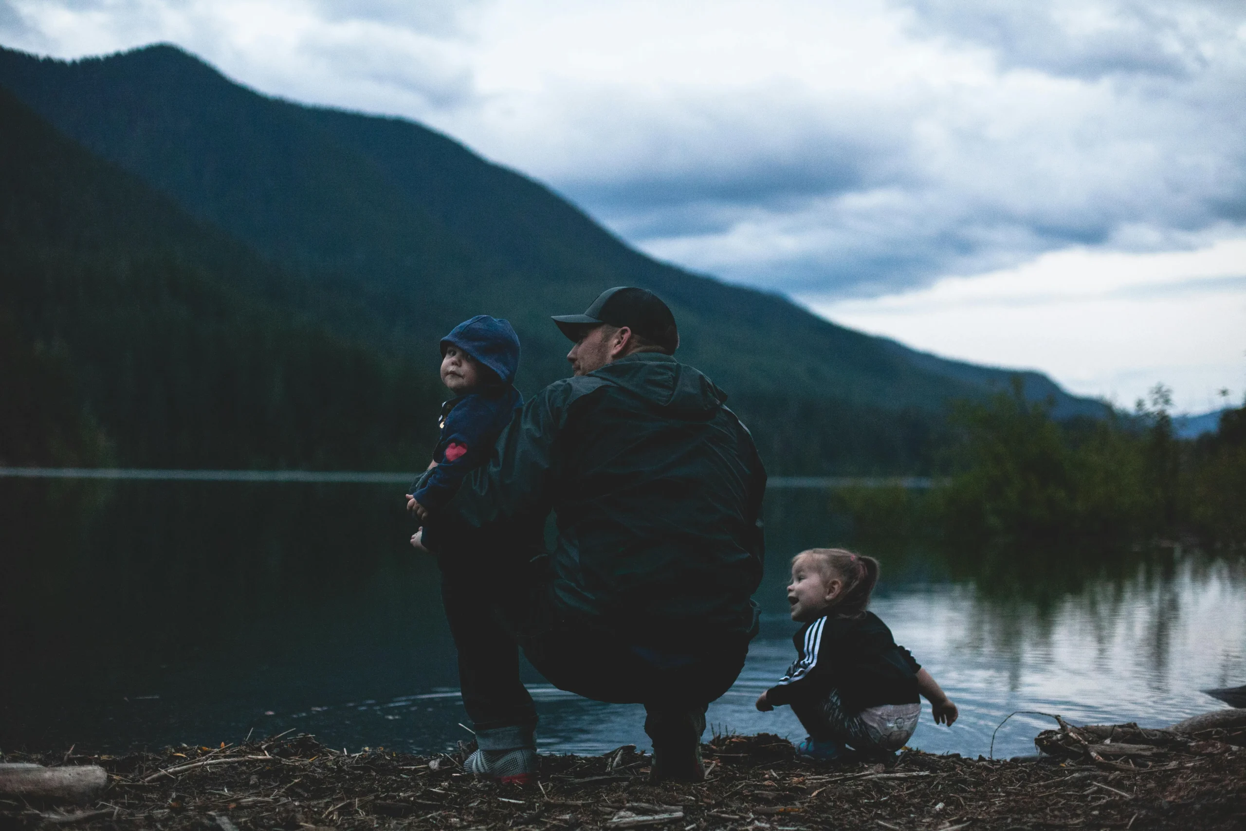 A father and his two young children looking over a beautiful lake with mountains in the distance