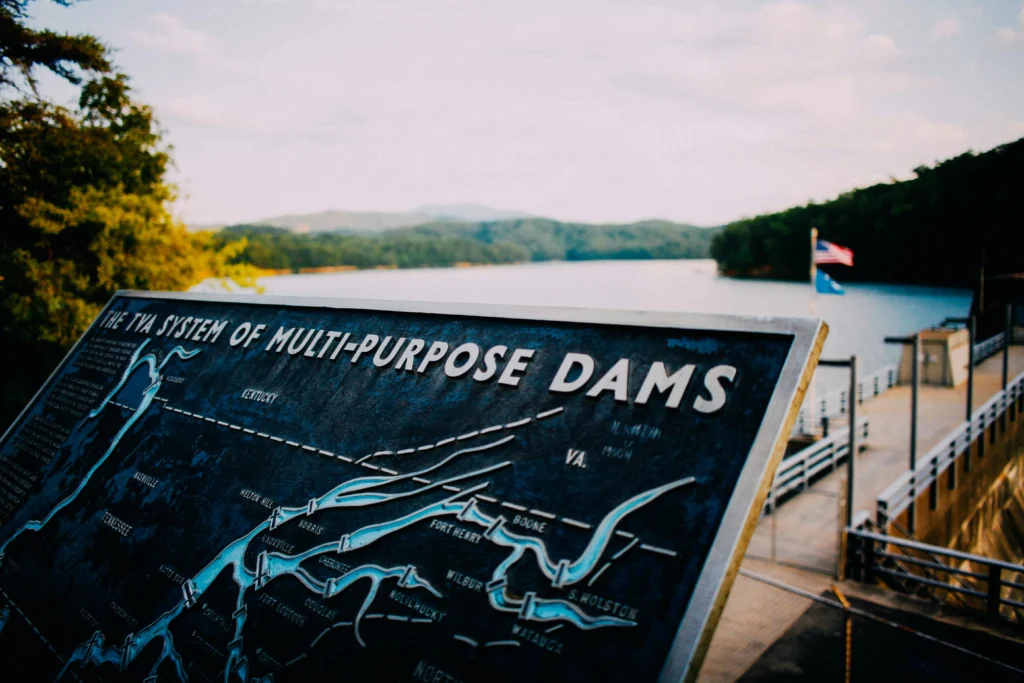 Close-up of a TVA system of multi-purpose dams sign showing river systems, with a lake and forested hills in the background under a cloudy sky.