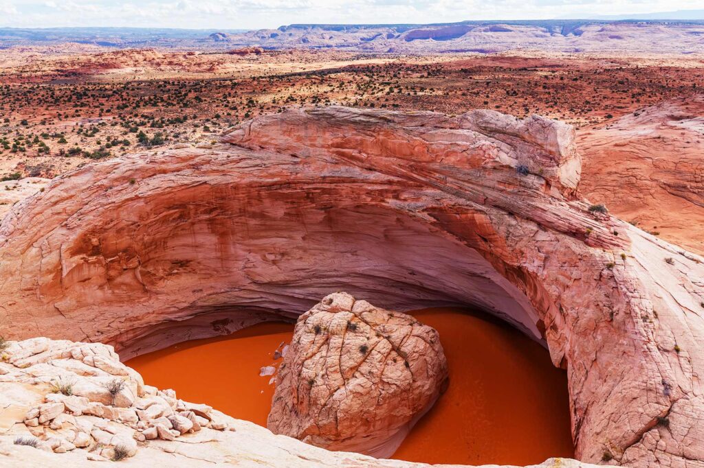 Overhead view of the Cosmic Ashtray, a massive rock bowl with a giant boulder in the center, surrounded by red desert terrain.