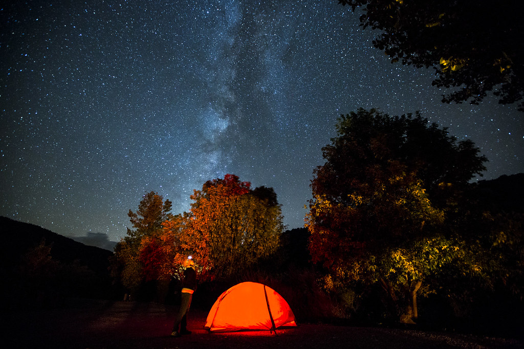 Milky Way stretching across a star-filled night sky above a glowing tent and silhouetted trees in Grand Staircase–Escalante National Monument.