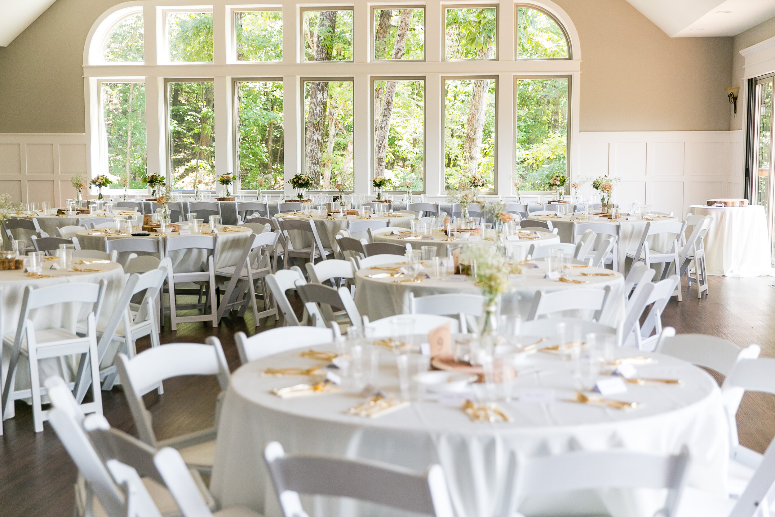 Elegant indoor wedding reception setup at Gardens in the Gorge featuring white round tables, white chairs, soft floral centerpieces, and large windows with forest views.