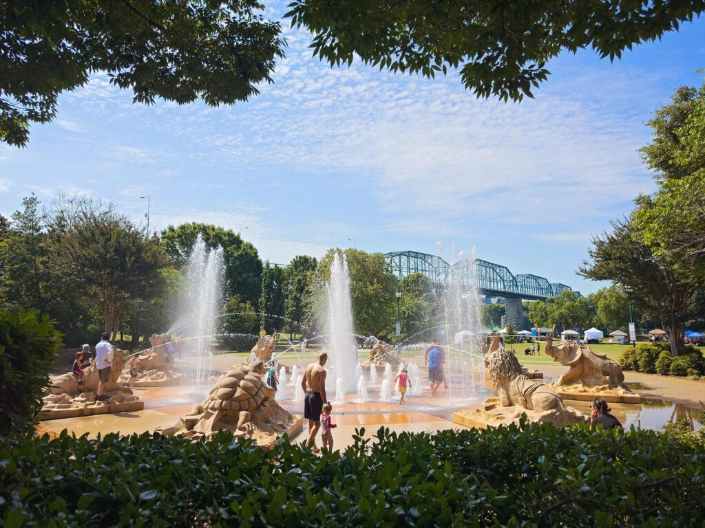 Children and families play in the interactive animal-themed fountain at Coolidge Park in Chattanooga, Tennessee, with large stone sculptures and water jets spraying under a sunny blue sky.