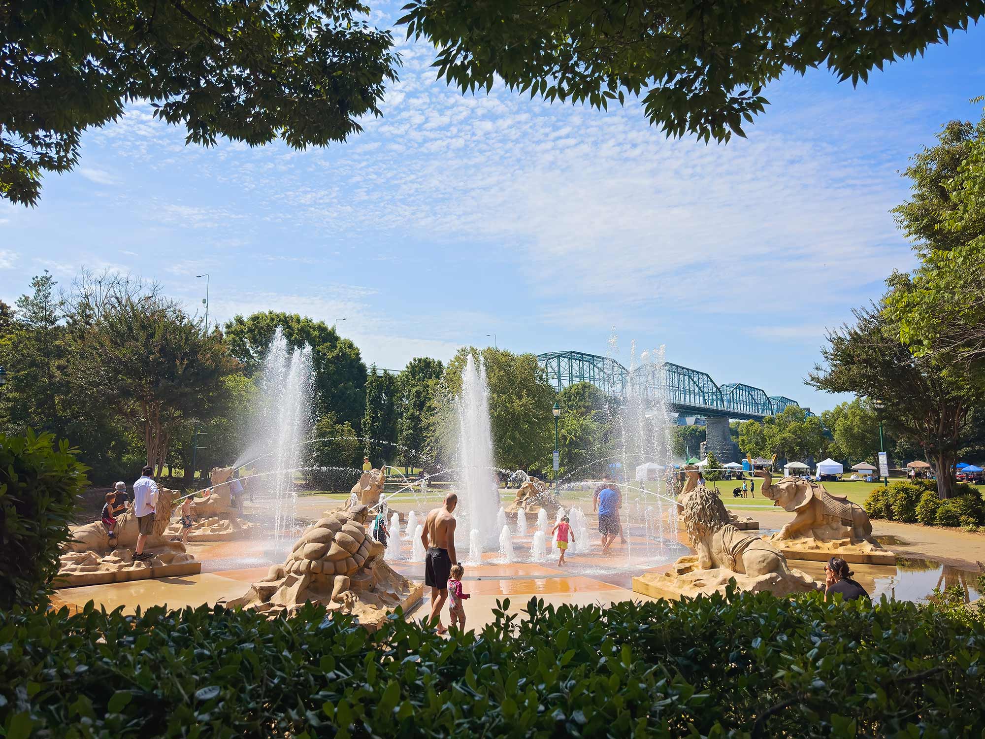 Children and families play in the interactive animal-themed fountain at Coolidge Park in Chattanooga, Tennessee, with large stone sculptures and water jets spraying under a sunny blue sky.