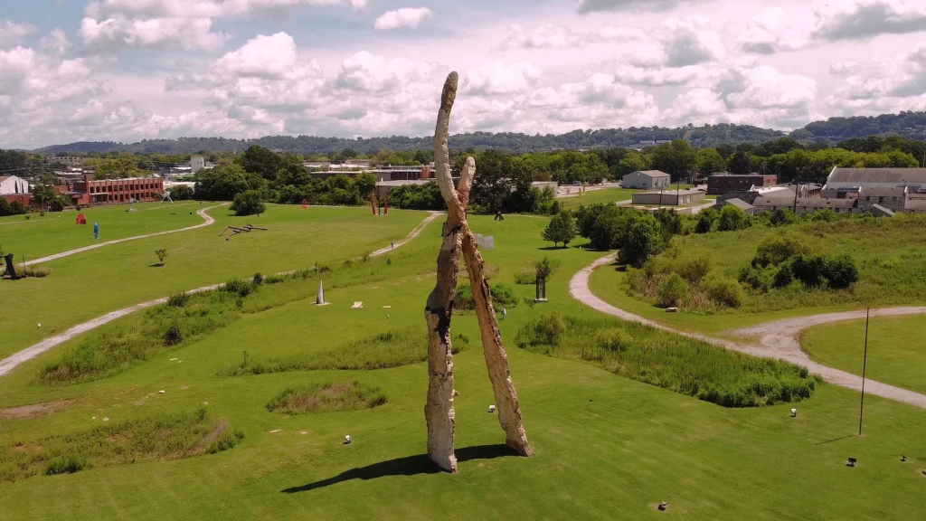 Wide view of Sculpture Fields at Montague Park in Chattanooga, Tennessee. A large abstract stone sculpture stands prominently in a grassy field surrounded by walking paths.