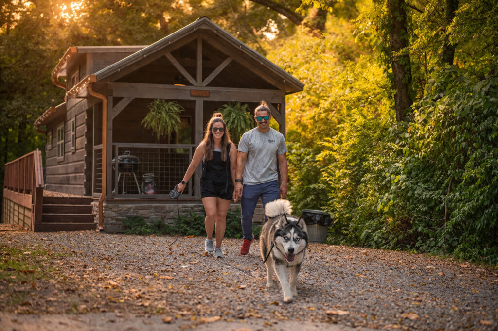 Couple walking a husky in front of a rustic pet-friendly cabin at Wauhatchie Woodlands in Chattanooga, Tennessee, surrounded by trees at sunset.