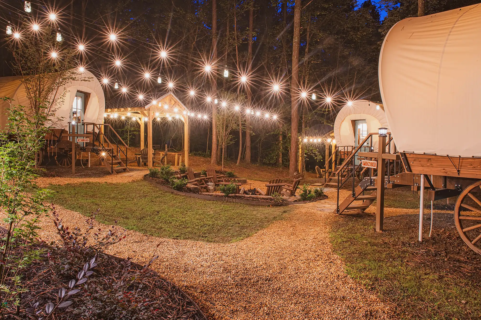 Glamping site with covered wagon cabins lit by string lights in a wooded setting