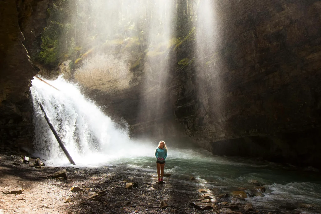 Girl standing beneath a waterfall in Nantahala National Forest, North Carolina, surrounded by rocky canyon walls and misty sunlight