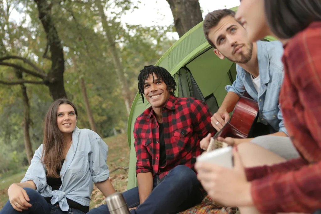 A group of smiling friends sitting around a campsite, with one man holding a guitar